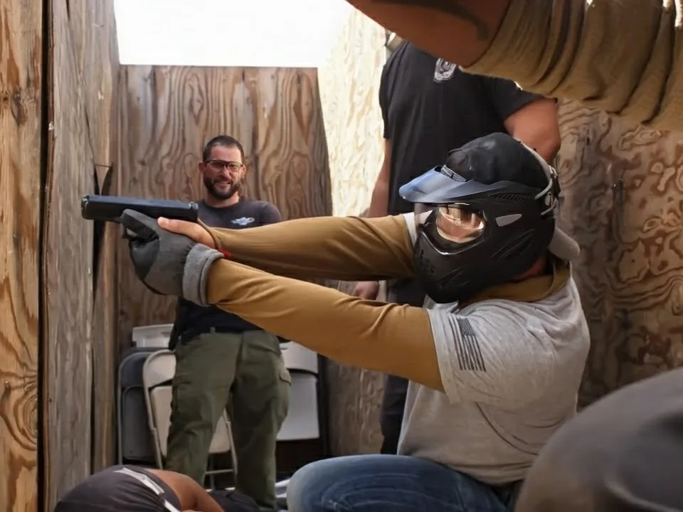 Student in a protective helmet aiming a training pistol inside a plywood shoot house while an instructor watches